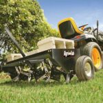 A lawn tractor towing an Agri-Fab spike aerator across a green lawn, with cinder blocks placed on top of the aerator for added weight to help penetrate the soil. The background includes green bushes and a blue sky.