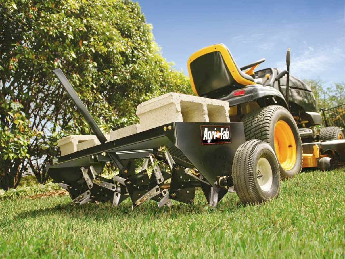 A lawn tractor towing an Agri-Fab spike aerator across a green lawn, with cinder blocks placed on top of the aerator for added weight to help penetrate the soil. The background includes green bushes and a blue sky.