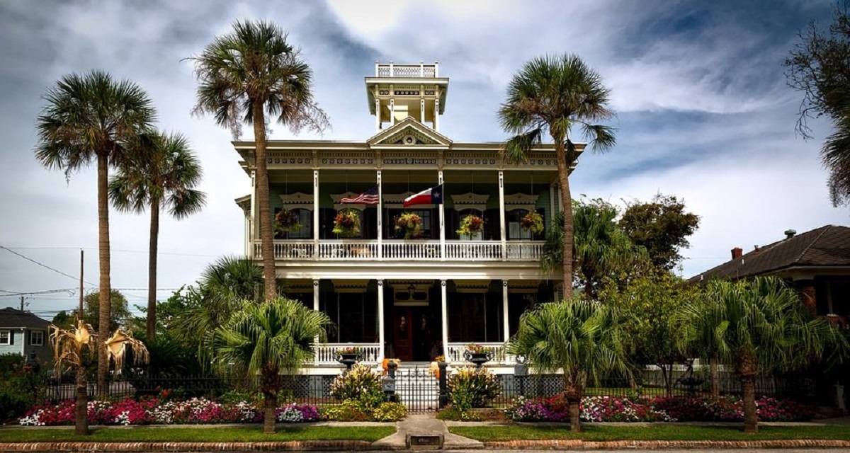 A two-story home with the American and Texan flags flying, surrounded by tall and short palm trees