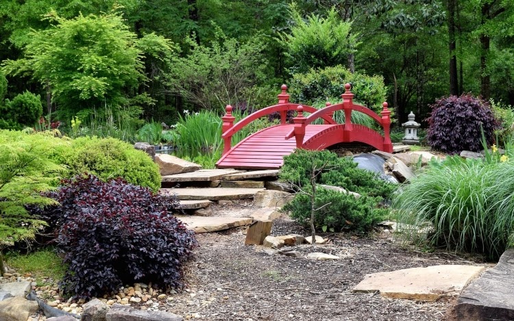 Red-colored wooden bridge overtop water feature, surrounded by stone pavers and various plants