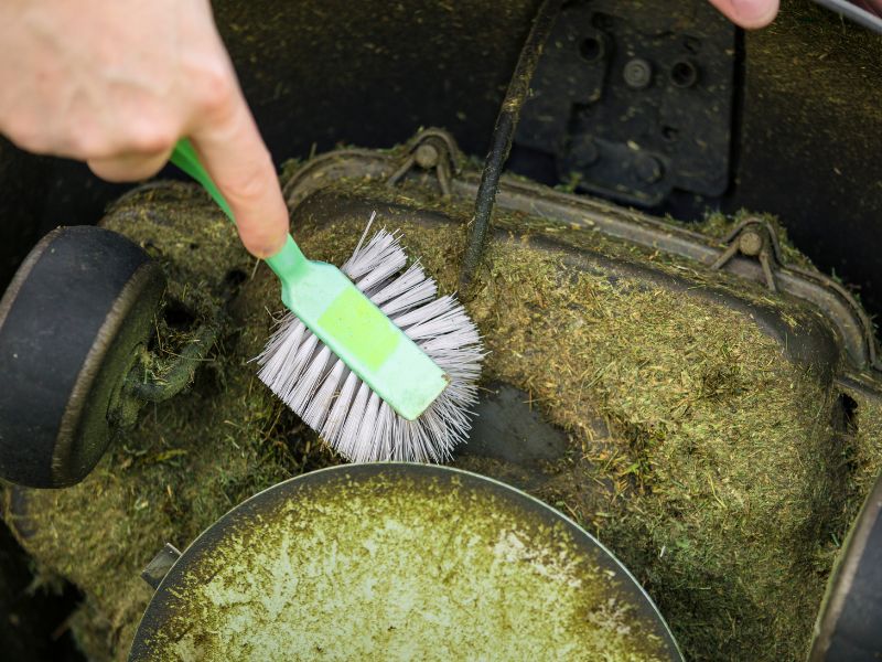 Close up view of man hand brushing off layer of wet grass stuck under automated lawnmower, maintenance concept.